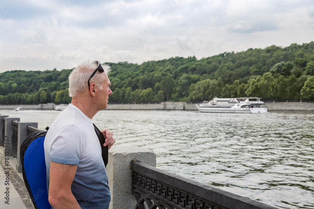 Fototapeta premium A sporty gray-haired man with a blue sports bag, wearing a T-shirt, stands on the river embankment on a cloudy day
