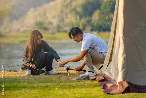 Happy couple camping in forest near lake, Asian man setting up camping tent outdoors, hammering the tent peg to the ground at the campsite while Woman playing with cat.