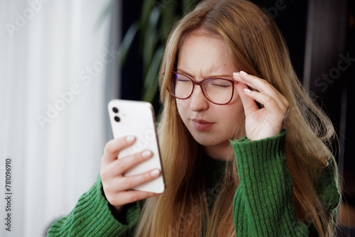 girl squinting with poor eyesight wearing glasses looking at a mobile phone at the table, concept of poor eyesight