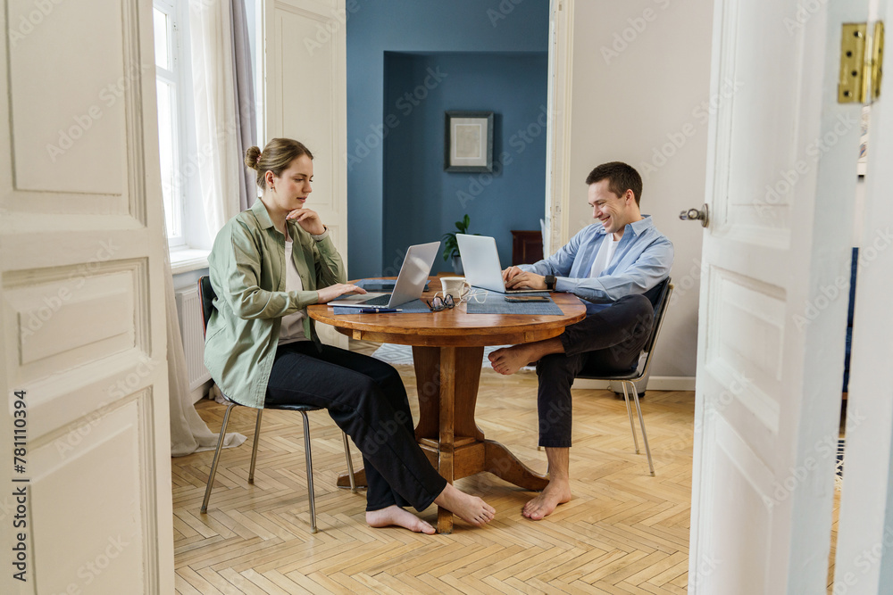 A barefoot couple, dressed in business casual attire, work side by side on their laptops at a wooden table, their casual posture reflecting the comfort of a home office. A barefoot couple, dressed in business casual attire, work side by side on their laptops at a wooden table, their casual posture reflecting the comfort of a home office.