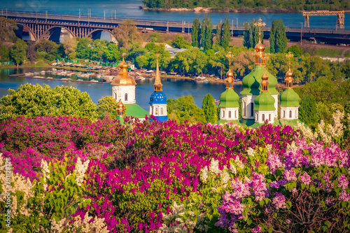 Golden domes of Kiev Pechersk Lavra, Ukraine, Europe. Blooming lilac trees at springtime in Orthodox Monastery. Stunning cityscape of Kiev and Dnipro river. Traveling concept background. photo