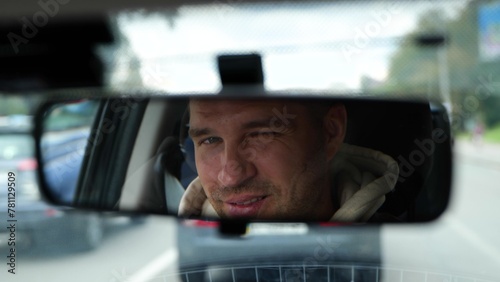 Happy young Caucasian male driving a car in city looking and winking in rearview mirror. Smiling handsome man face reflected in car mirror. Traveling in vehicle. Close up © Nataliya