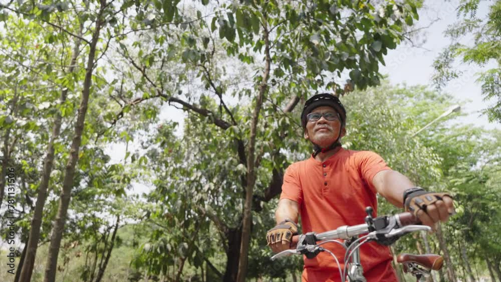 Active Elderly Man in Orange Cycling Gear Looking Up While Riding Bike in a Verdant Park Active Elderly Man in Orange Cycling Gear Looking Up While Riding Bike in a Verdant Park