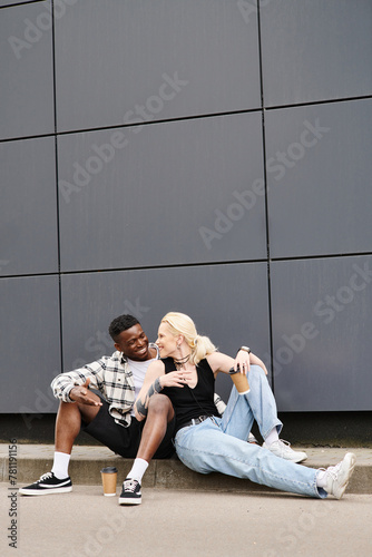 A happy multicultural couple sitting beside each other on the ground near a grey urban building, sharing a quiet moment.