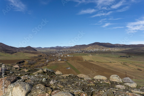 View of the village and mountains from the Medieval fortress on a rock. Stone wall and agricultural fields.  Bright blue sky with clouds.  Kveshi fortress. Georgia