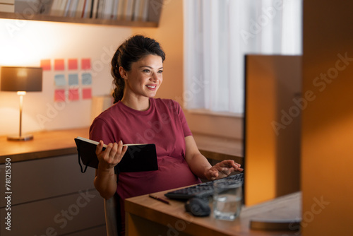 A cheerful mid adult pregnant woman entering financial data in her computer and working from home