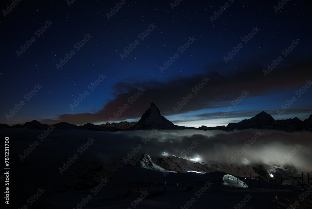 Breathtaking night scene at Zermatt ski resort, Matterhorn mountain silhouetted against starry sky. Tranquil with hint of human activity, serene natural beauty.