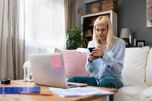 Taking a break. Young freelance business woman respond on text messages on her smartphone in pause of work on laptop computer. Online working at home, female online dating during the day.