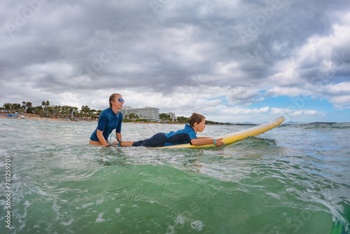 Happy boy learns to surf - young surfer learn to ride on surfboard with surf instructor on sea waves. Active family lifestyle, kids outdoor water sport lessons. © Igor