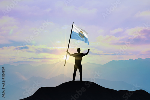 San Marino flag being waved by a man celebrating success at the top of a mountain against sunset or sunrise. San Marino flag for Independence Day.