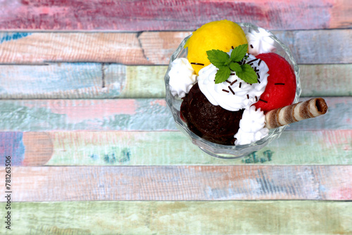 Top view on sundea ice cream - three kinds of ice cream in glass cup for ice cream with whipped cream, mint and biscuit stick on color table, hot summmer sunny day, space for text photo