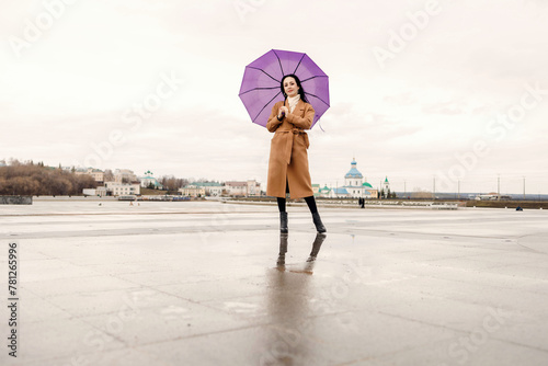A girl in a light brown coat and a light scarf under a purple umbrella shot on a cloudy April evening against the background of Cheboksary photo