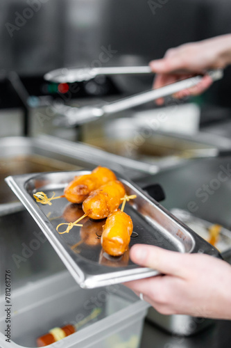 Professional chef preparing glazed appetizers in a kitchen. Golden brown, glossy appetizers are in focus on a stainless steel tray, with blurred kitchen equipment in the background