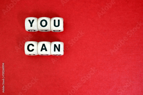 Inspirational mantra You Can spelt with wooden word dice above a red cardboard background.