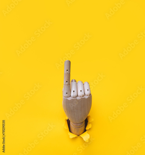 A wooden hand protrudes from a torn hole in yellow paper and points its index finger upward. Concept of direction, adjustment and assistance. Mannequin, robot.