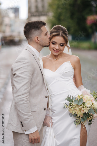 Portrait happy newlyweds walking outdoors. Handsome groom in light colour suit and bride in elegant wedding dress
