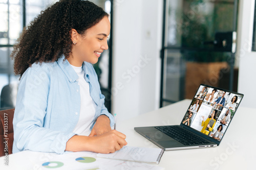 E-learning, online webinar. Brazilian or hispanic woman, sits at workplace in the office in front of laptop, having online video call with multiracial people, listening online lecture, takes notes
