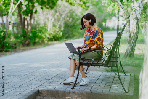 A cheerful woman works on her laptop while sitting on a park bench surrounded by birch trees. Casual businesswoman smiling as she successfully multitasks in urban park