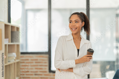Young Black businesswoman smiling confidently while holding a takeaway coffee cup in a light modern office, representing success, aspirations, and a positive outlook at work Free Download Young Black businesswoman smiling confidently while holding a takeaway coffee cup in a light modern office, representing success, aspirations, and a positive outlook at work - GFXLoad