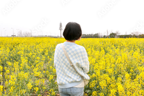 Young fashionable asian woman with her back to us, in the middle of a rapeseed (Brassica napus) field surrounded by bright colourful yellow flowers, enjoying the spring summer sunlight