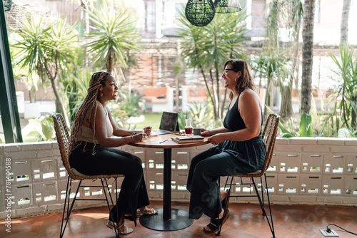 Two women enjoy collaborative work in tropical café. woman with braids smiles as she listens, while her companion gestures during conversation. laptop and note-taking indicate productive atmosphere
