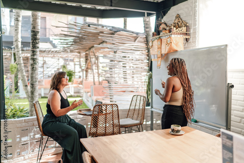 Two women, one with long braided hair and another with glasses interacting with excitement at sunlit cafe, surrounded by greenery. dynamic discussion brainstorm or business presentation