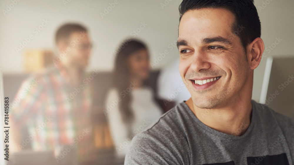 Young businessman with arms crossed in a office, smiles while his three diverse colleagues are in the background talking together Young businessman with arms crossed in a office, smiles while his three diverse colleagues are in the background talking together