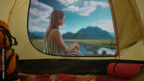 Female resting on campsite relaxing near lake. Young woman sitting outside the tent at daylight in the mountains lake coast, listening music in headphones.