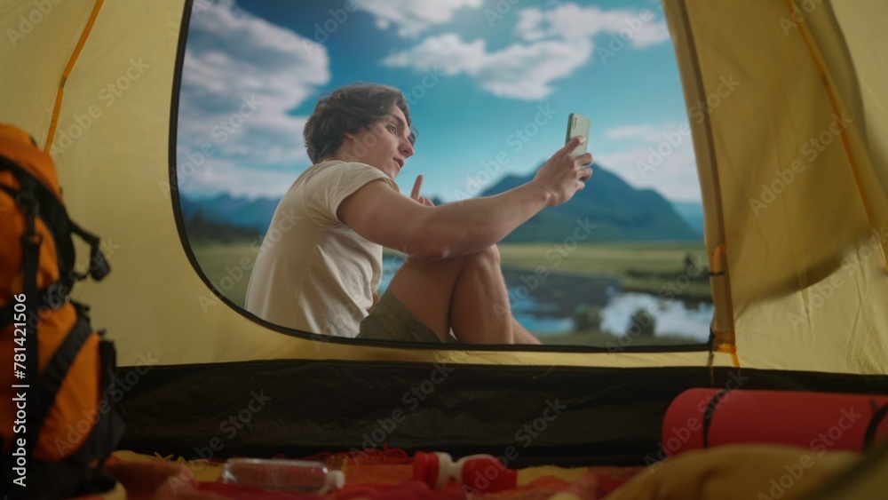 Male resting on campsite relaxing near lake. Young man sitting outside the tent at daylight in the mountains lake coast, taking selfie pictures on smartphone.