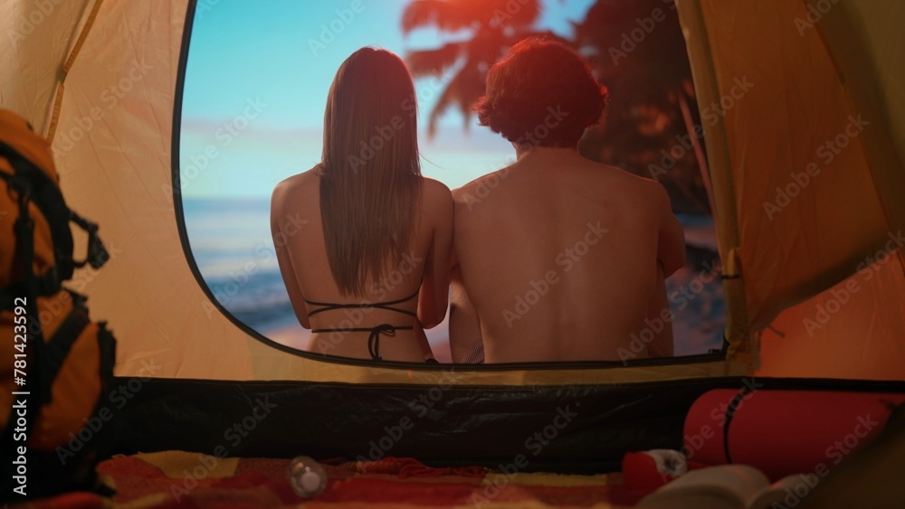 Couple resting on campsite relaxing near ocean. Young man and woman sitting outside the tent at sunset on the beach looking at sea, vacation outdoors.