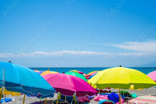 A beach scene with many colorful umbrellas and a clear blue sky. Scene is cheerful and inviting, as the beachgoers are enjoying the sunny day