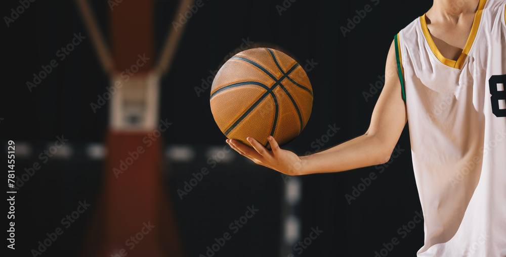 Young basketball player holds classic ball. Basket in background. Junior level basketball player holding game ball. Basketball training session for kids