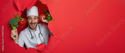 Charismatic male chef with fresh greens and tomato smiling through a torn hole in a red paper background, stethoscope visible © Fxquadro