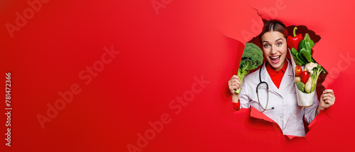 Happy female chef with a stethoscope bursts through a red background, holding a variety of greens and vegetables © Fxquadro