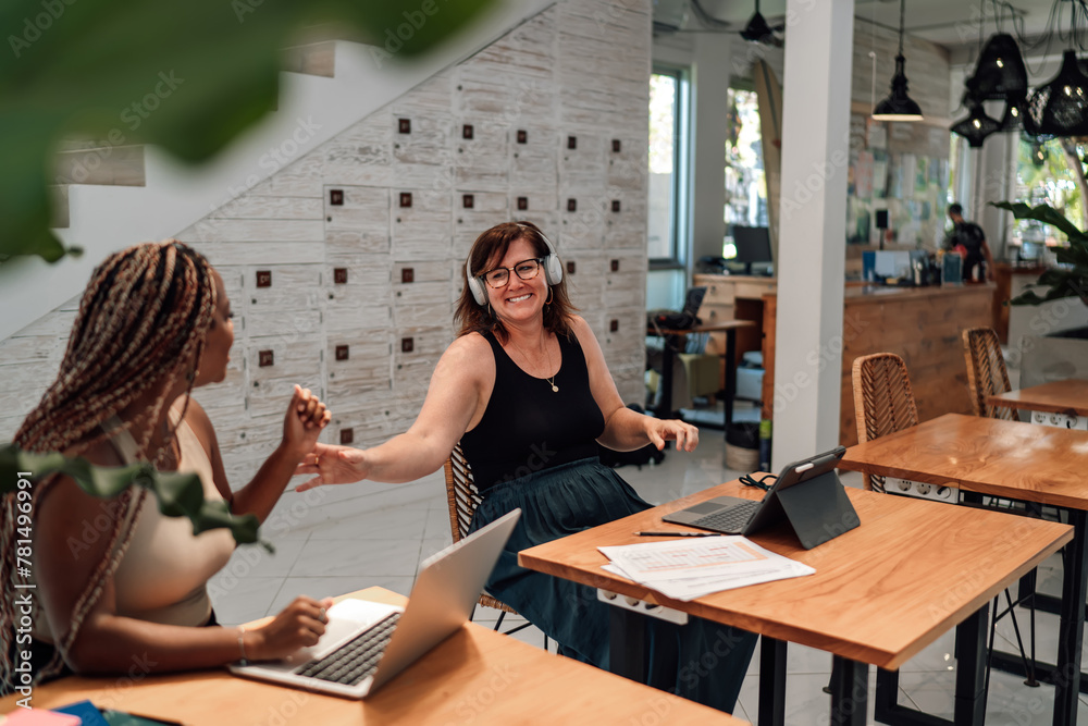 joyful interaction between two businesswomen at office table, one African-American with laptop, other Caucasian wearing headphones, in well-lit workspace, teamwork and positive corporate culture joyful interaction between two businesswomen at office table, one African-American with laptop, other Caucasian wearing headphones, in well-lit workspace, teamwork and positive corporate culture