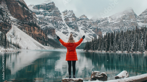 A woman in a red jacket is standing on a rock overlooking a serene lake