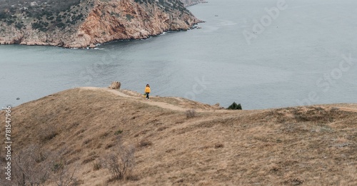 Woman stands on cliff overlooking ocean, cloudy day provides soft lighting. She's wearing a yellow sweater wearing dark pants. Landscape features rugged terrain, calm waters.