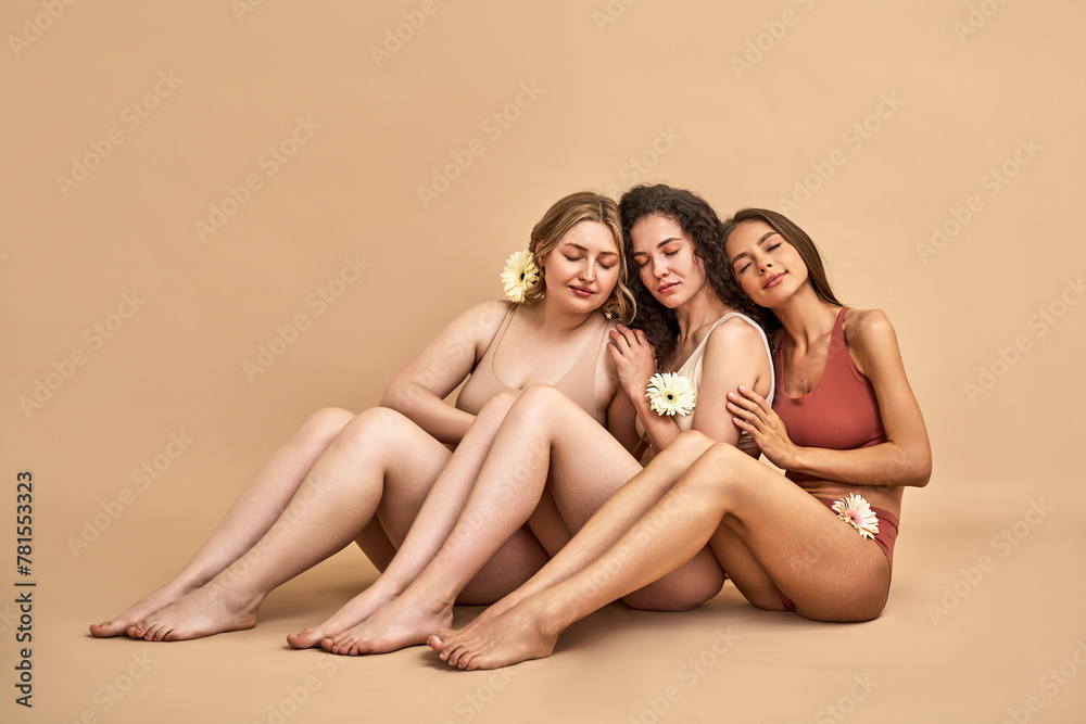 Self love. Tender young women with different body and skin types sitting over beige background in lingerie, keeping eyes closed and holding gerberas. Group solidarity and health care concept. Self love. Tender young women with different body and skin types sitting over beige background in lingerie, keeping eyes closed and holding gerberas. Group solidarity and health care concept.