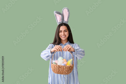 Beautiful young happy woman in bunny ears holding basket with Easter painted eggs on green background