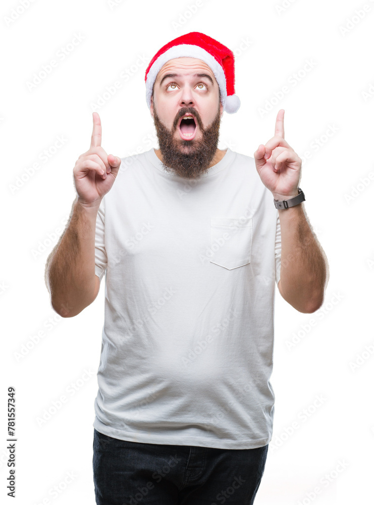 Young caucasian hipster man wearing christmas hat over isolated background amazed and surprised looking up and pointing with fingers and raised arms. Young caucasian hipster man wearing christmas hat over isolated background amazed and surprised looking up and pointing with fingers and raised arms.