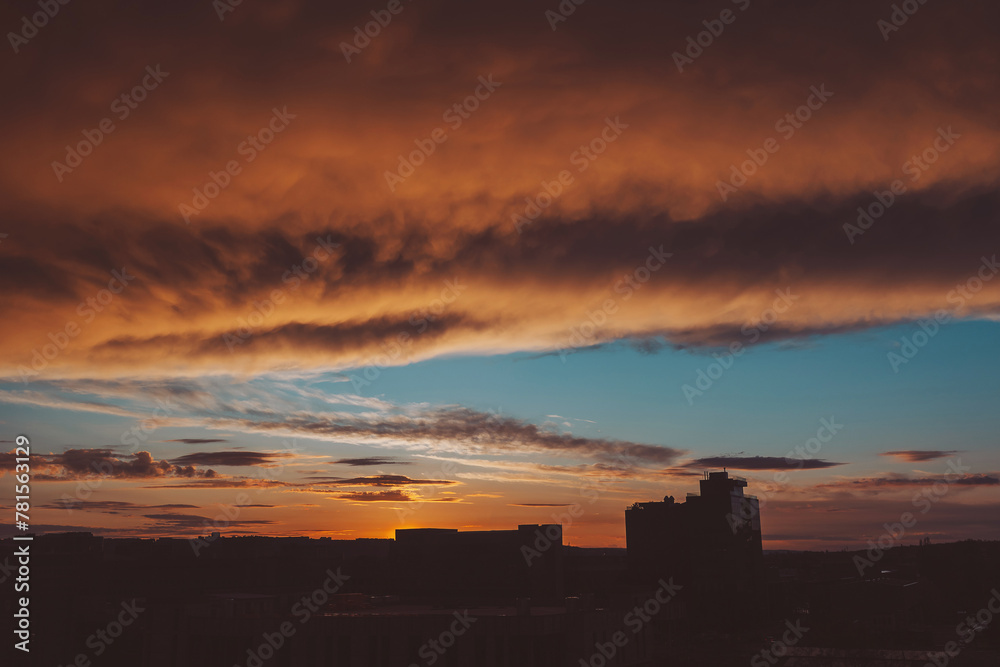 Fototapeta premium Sunrise clouds, some dark, some light up by the sun, over high rise buildings. Cityscape with wonderful fiery dawn. Amazing dramatic cloudy sky. Atmospheric background of sunrise in overcast weather.