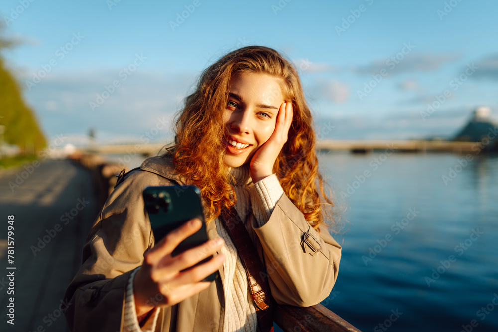 Selfie time. Young woman holding mobile phone taking selfie photo using smartphone camera standing on waterfront. Lifestyle, travel, tourism, vacation, technology, weekend.
