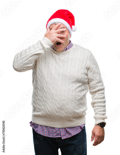 Handsome senior man wearing christmas hat over isolated background peeking in shock covering face and eyes with hand, looking through fingers with embarrassed expression.