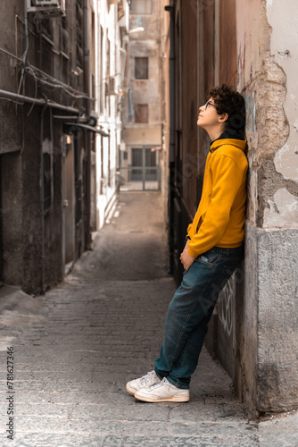 A thoughtful teenager in casual style leans against a textured wall in an alley, embodying urban solitude and contemplation amidst the cityscape.