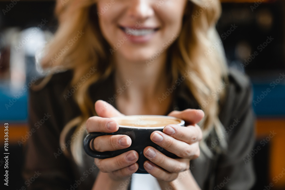 Fototapeta premium Focused shot. Young caucasian beautiful woman holding cup mug of hot beverage coffee tea enjoying aroma while having breakfast brunch in cafe restaurant