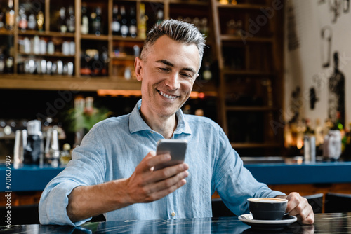 Smiling confident caucasian mature businessman freelancer boss using cellphone for surfing net, social media, e-banking, working remotely while drinking coffee in cafe restaurant