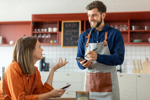Handsome smiling waiter using application on digital tablet, taking order of young female client in pastry shop of cafe