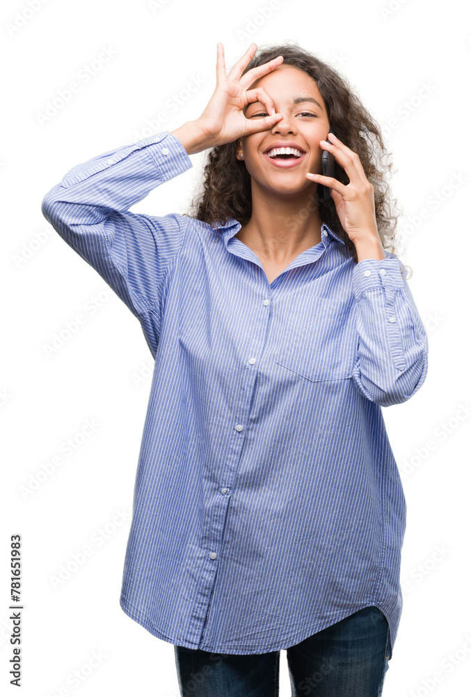 Young hispanic woman using smartphone with happy face smiling doing ok sign with hand on eye looking through fingers Young hispanic woman using smartphone with happy face smiling doing ok sign with hand on eye looking through fingers