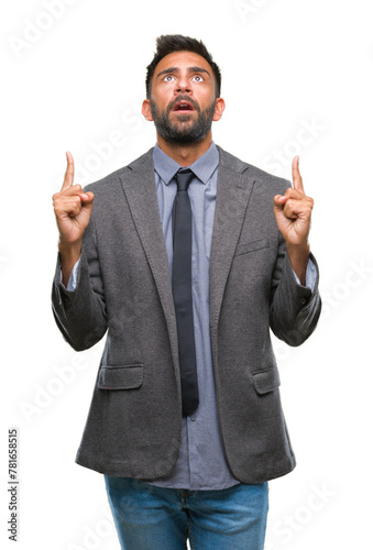 Adult hispanic business man over isolated background amazed and surprised looking up and pointing with fingers and raised arms.
