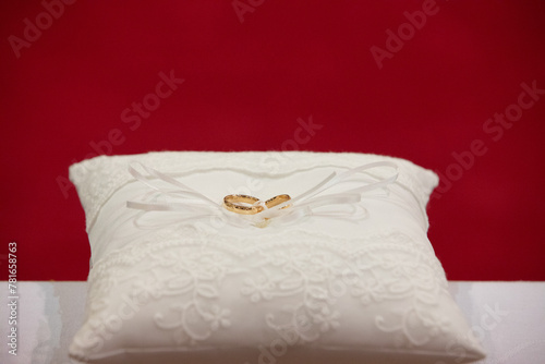 golden wedding rings on white embroidered pillow against red background